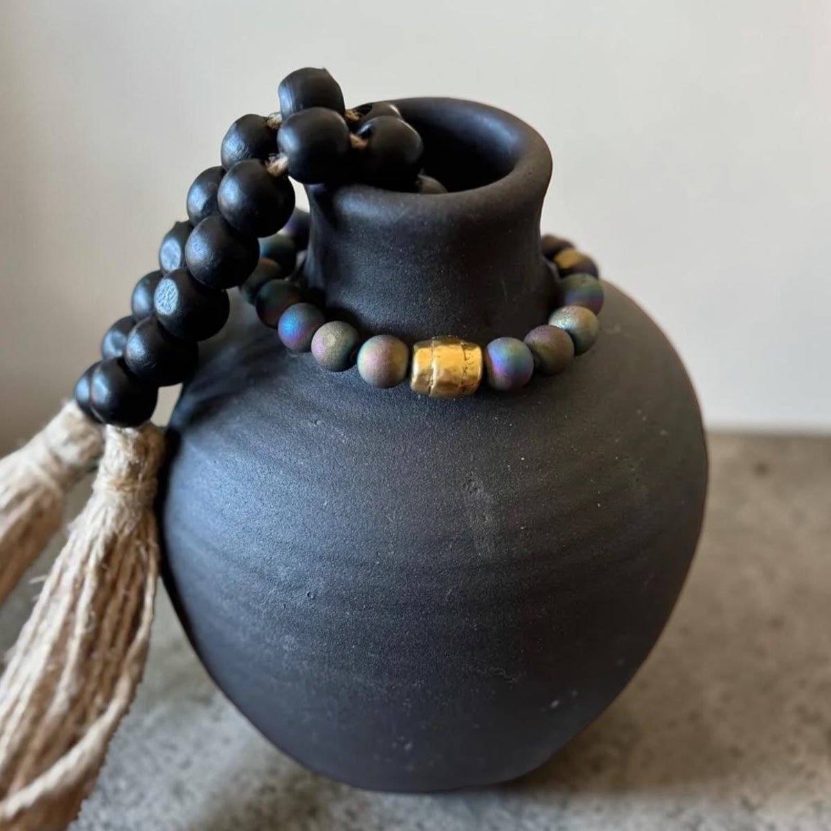 Black wooden prayer beads with a tassel and a small gold accent bead, draped over a matte black ceramic vase against a neutral background.