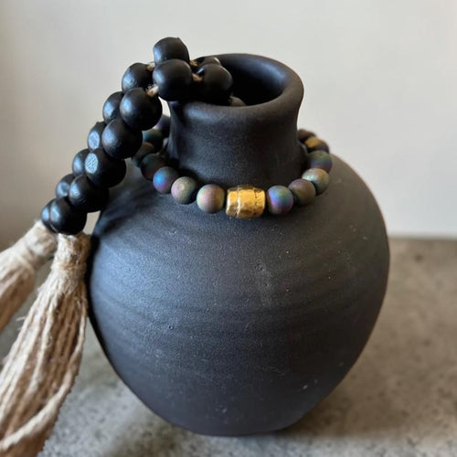 Black wooden prayer beads with a tassel and a small gold accent bead, draped over a matte black ceramic vase against a neutral background.