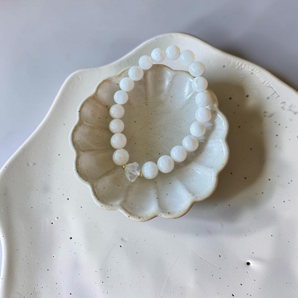 A beaded bracelet made of matte white round beads with a small clear crystal accent, displayed inside a scalloped ceramic dish on a speckled white plate.