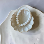 A beaded bracelet made of matte white round beads with a small clear crystal accent, displayed inside a scalloped ceramic dish on a speckled white plate.