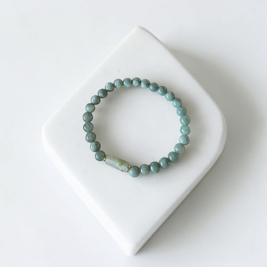 A beaded stretch bracelet in a muted sage green color, displayed on a white ceramic stand against a clean white background.