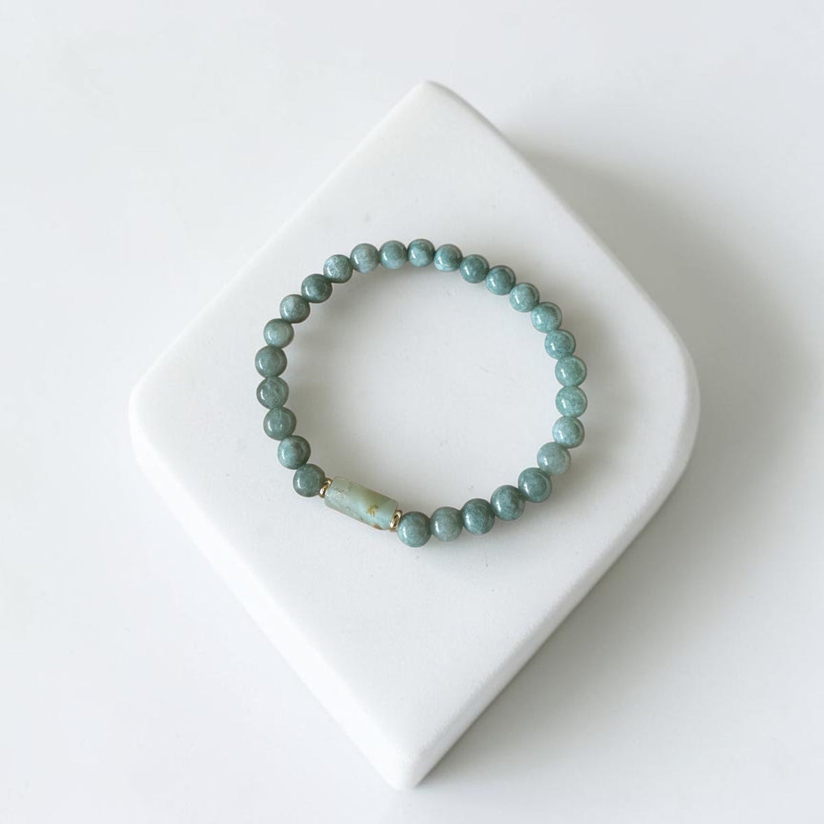 A beaded stretch bracelet in a muted sage green color, displayed on a white ceramic stand against a clean white background.