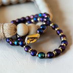 A close-up of a beaded bracelet with iridescent dark purple and blue beads, small gold spacer beads, a gold-toned leaf charm, and a few natural cream wooden beads, resting on a light textured surface.