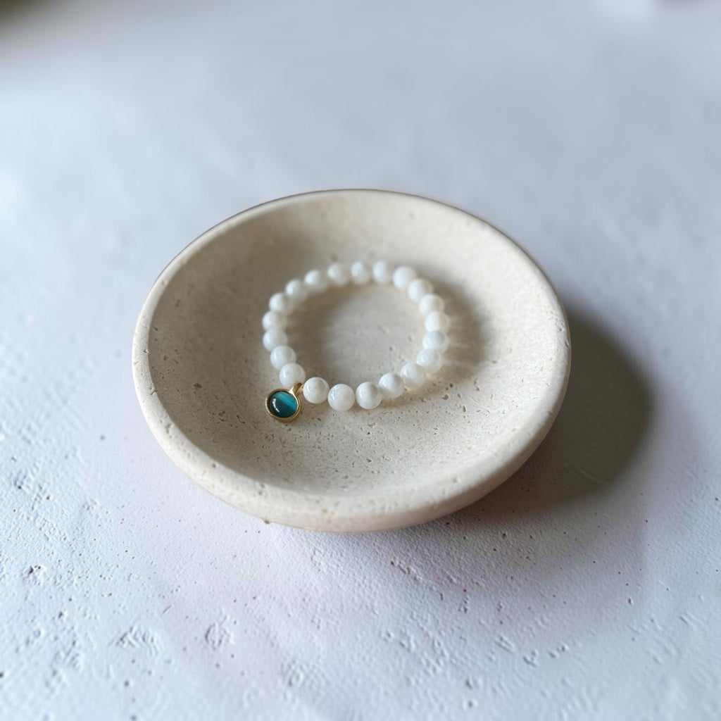A beaded stretch bracelet with white round beads and a small blue-green stone charm, displayed inside a small speckled ceramic dish on a textured white surface.