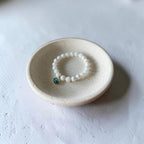 A beaded stretch bracelet with white round beads and a small blue-green stone charm, displayed inside a small speckled ceramic dish on a textured white surface.