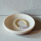 A delicate beaded stretch bracelet with gold-toned round beads and small lavender-purple translucent beads, displayed inside a shallow speckled ceramic dish on a neutral surface.