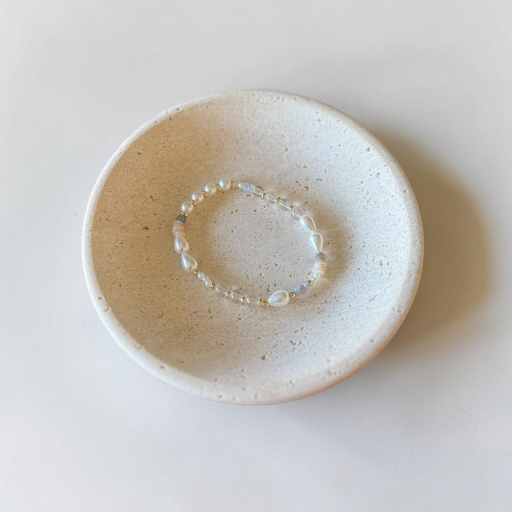 A delicate beaded bracelet with iridescent white beads rests inside a small, round, speckled ceramic dish on a clean white surface.