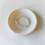 A delicate beaded bracelet with iridescent white beads rests inside a small, round, speckled ceramic dish on a clean white surface.