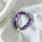A beaded stretch bracelet featuring round purple and violet beads, with a single white baroque pearl accent and small gold spacer beads, displayed on a draped white satin fabric.