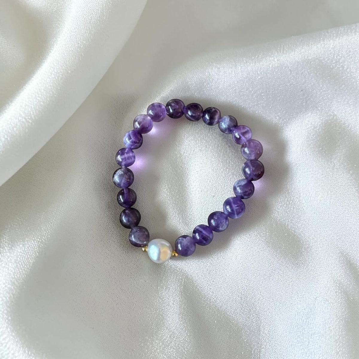 A beaded stretch bracelet featuring round purple and violet beads, with a single white baroque pearl accent and small gold spacer beads, displayed on a draped white satin fabric.