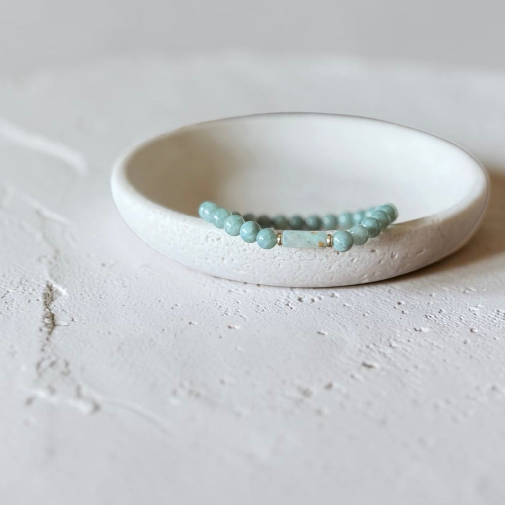 A beaded stretch bracelet in light dusty green blue, displayed on a small white ceramic dish against a textured light gray background.