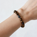 A beaded stretch bracelet worn on a person's wrist, featuring glossy brown and black tiger's eye beads with one dark polished oval bead, set against a plain light background.