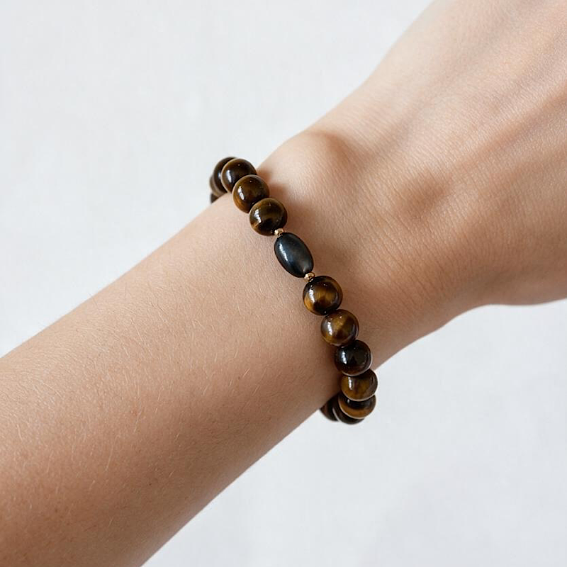A beaded stretch bracelet worn on a person's wrist, featuring glossy brown and black tiger's eye beads with one dark polished oval bead, set against a plain light background.