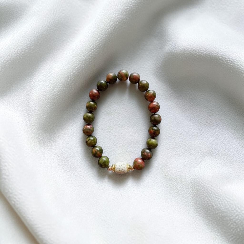 A beaded stretch bracelet made of round green and reddish-brown beads with a small white textured accent bead, displayed on a white fabric background.