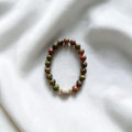 A beaded stretch bracelet made of round green and reddish-brown beads with a small white textured accent bead, displayed on a white fabric background.
