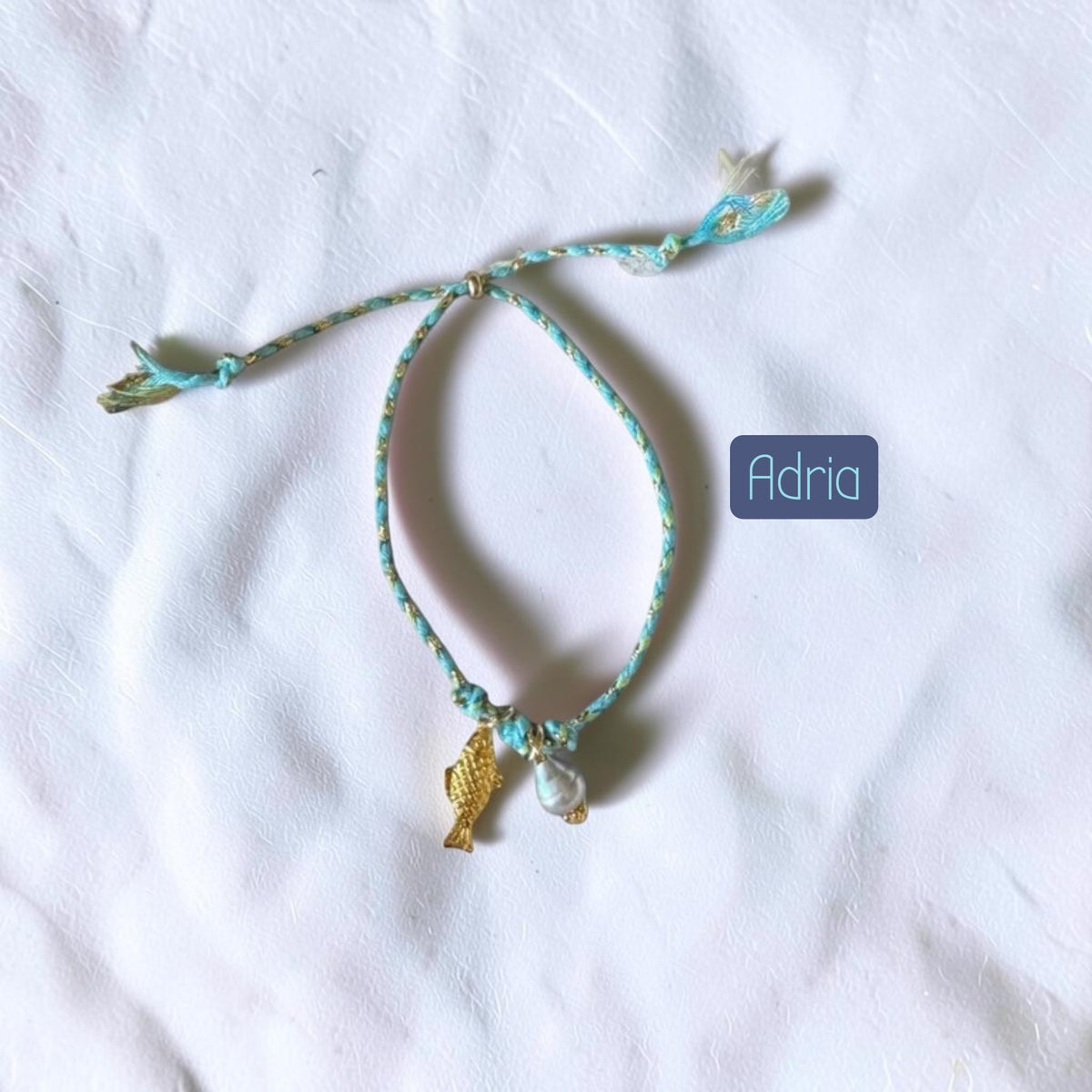 A handmade turquoise and white braided cord bracelet with a small gold fish charm and a white pearl bead, displayed on a white fabric background with a brand label reading 'Adria'.