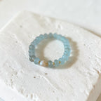 A beaded stretch bracelet featuring light blue translucent beads, a small white pearl, and tiny gold spacer beads, displayed on a textured white stone block.