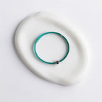 A thin teal-green beaded bracelet with a small iridescent bead and tiny gold-toned accents, displayed on a white ceramic dish against a clean white background.
