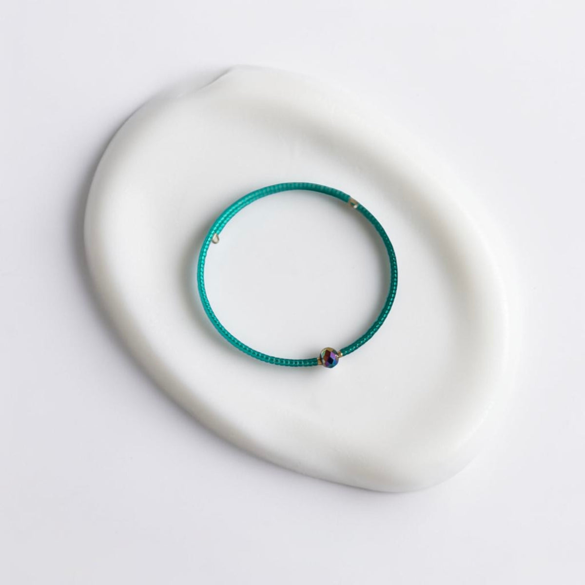 A thin teal-green beaded bracelet with a small iridescent bead and tiny gold-toned accents, displayed on a white ceramic dish against a clean white background.