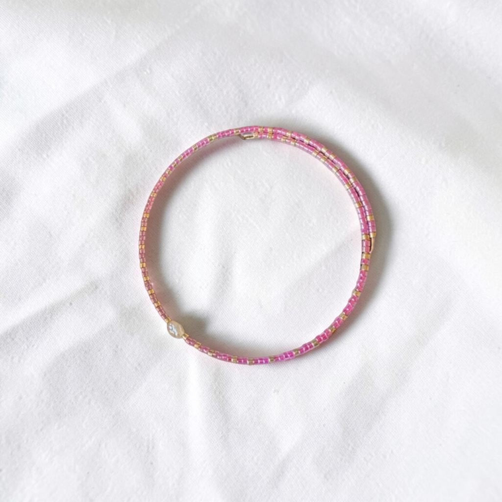A thin beaded bracelet in pink and gold, featuring a small white pearl accent, displayed on a textured white fabric background.