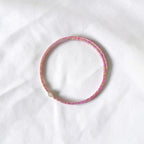 A thin beaded bracelet in pink and gold, featuring a small white pearl accent, displayed on a textured white fabric background.