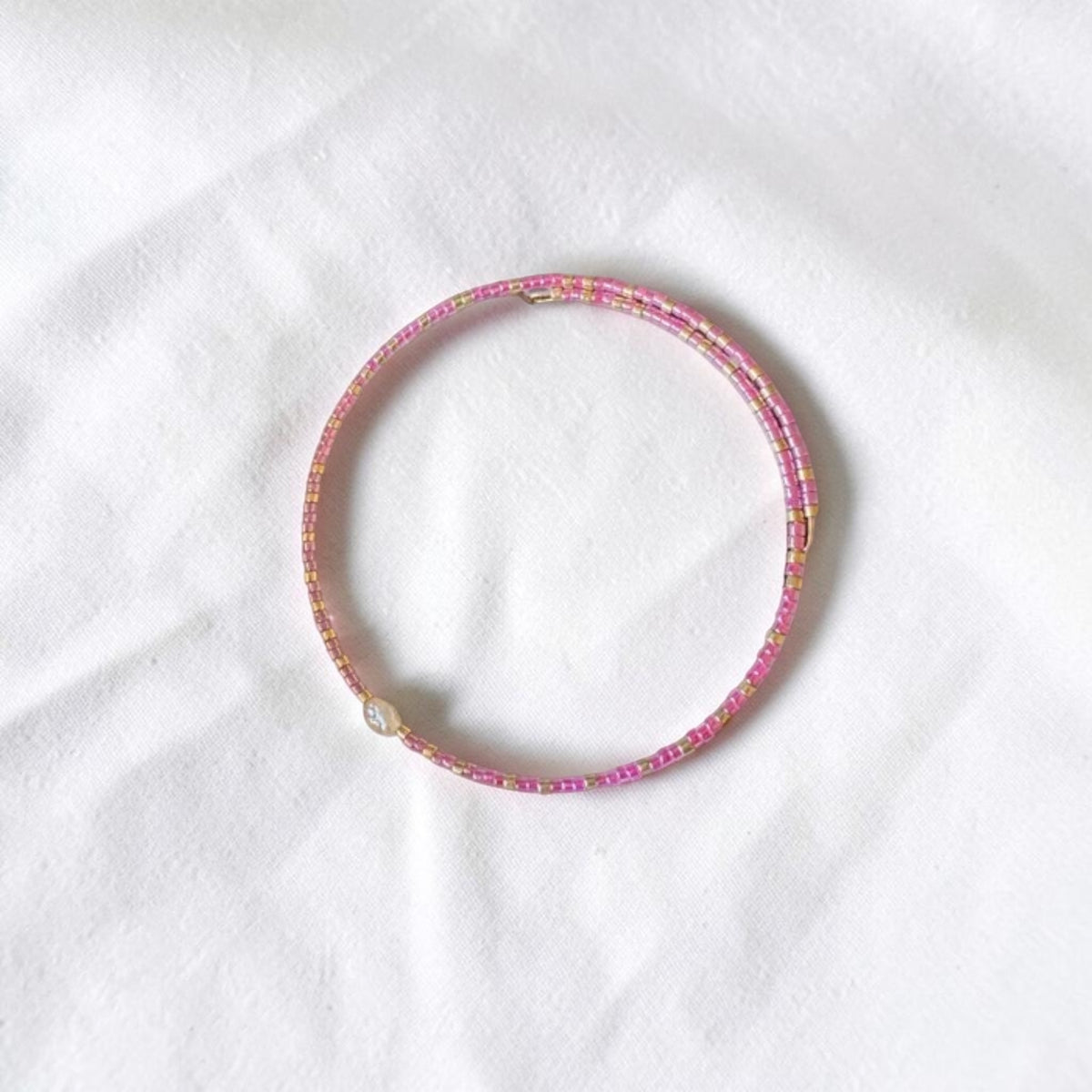 A thin beaded bracelet in pink and gold, featuring a small white pearl accent, displayed on a textured white fabric background.
