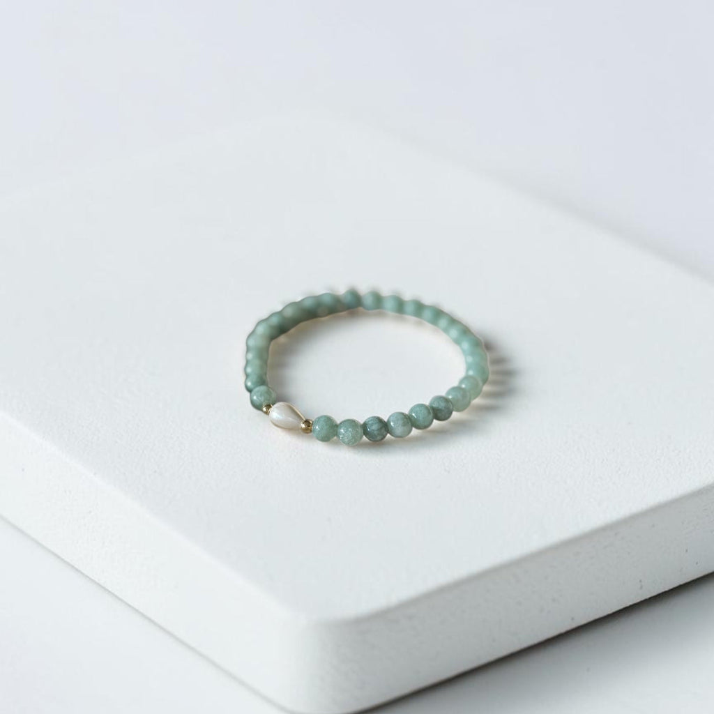 A beaded stretch bracelet with light green beads and a small gold accent, resting on a white textured rectangular block against a white background.