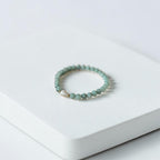 A beaded stretch bracelet with light green beads and a small gold accent, resting on a white textured rectangular block against a white background.