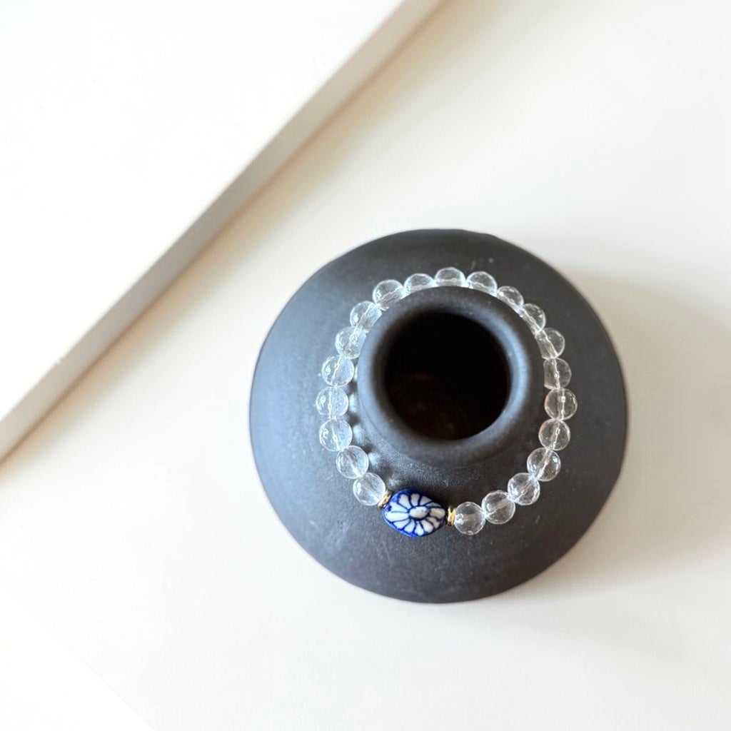 A small round black ceramic vase or candle holder with a central opening, styled with a clear beaded garland and a blue-and-white floral bead on a warm off-white surface.