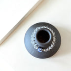 A small round black ceramic vase or candle holder with a central opening, styled with a clear beaded garland and a blue-and-white floral bead on a warm off-white surface.
