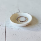 A beaded stretch ring with translucent brown and white beads rests inside a small, round, off-white ceramic dish on a distressed white wooden surface.