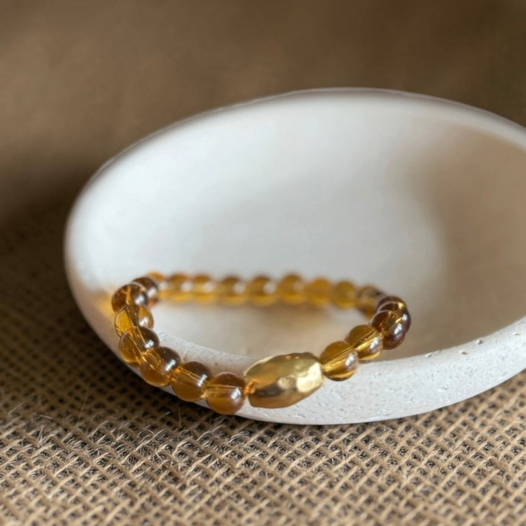 A beaded stretch bracelet made of translucent amber-yellow beads, displayed on a small white ceramic dish over a brown burlap surface.