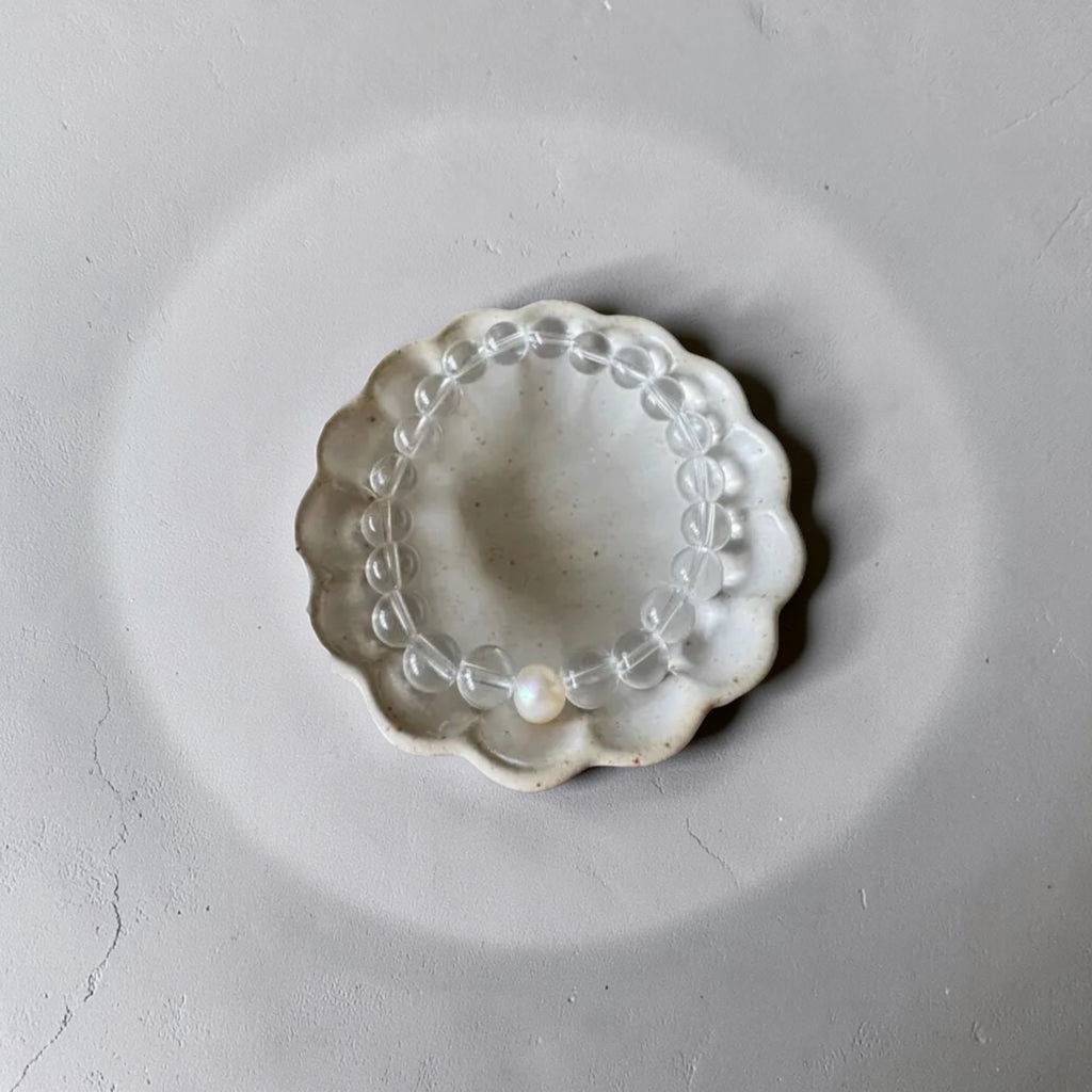 A clear beaded bracelet with a single white pearl bead, displayed inside a small scalloped off-white ceramic dish on a textured gray background.