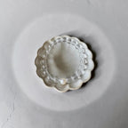 A clear beaded bracelet with a single white pearl bead, displayed inside a small scalloped off-white ceramic dish on a textured gray background.