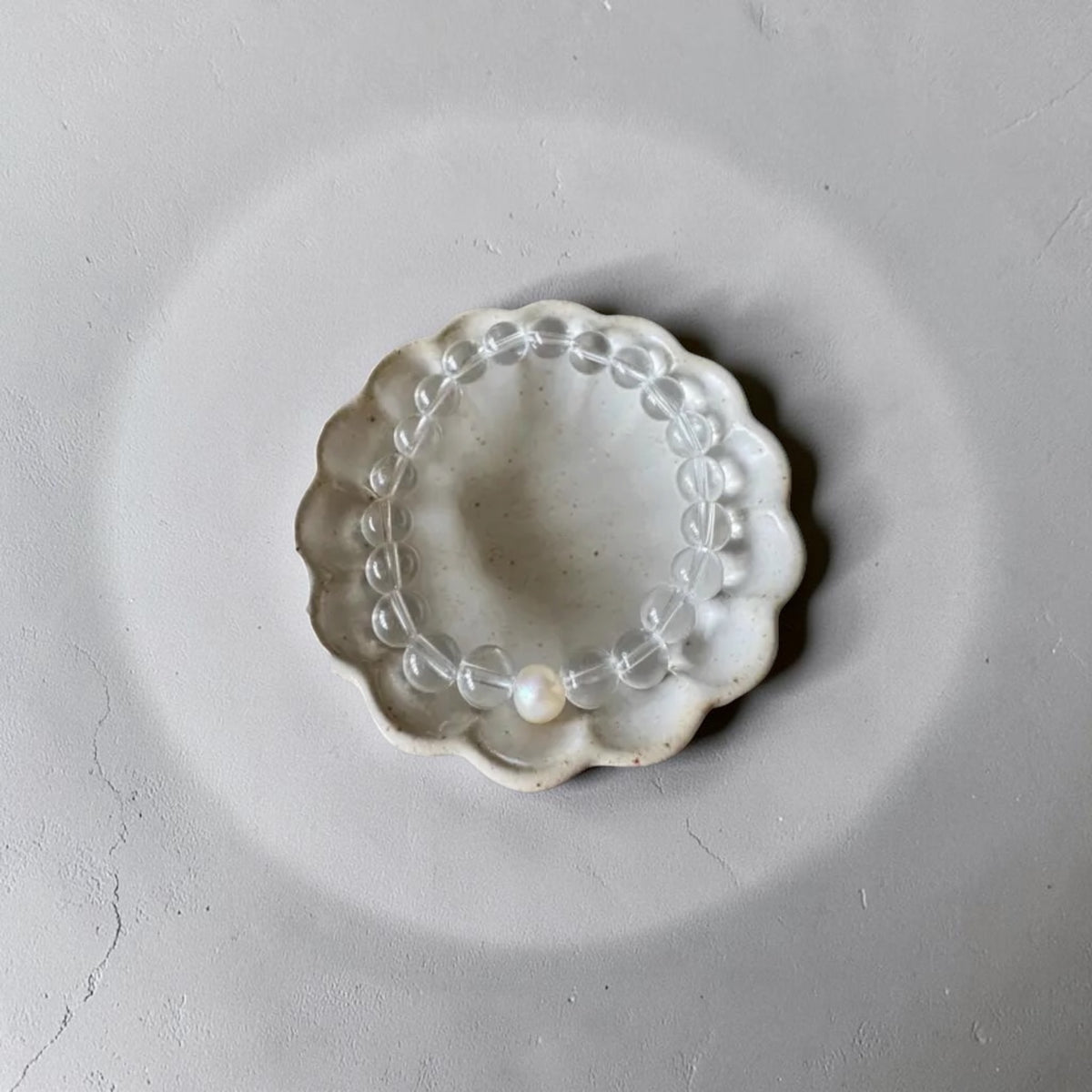 A clear beaded bracelet with a single white pearl bead, displayed inside a small scalloped off-white ceramic dish on a textured gray background.