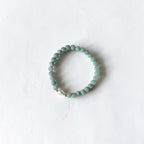 A beaded stretch bracelet made of small round light green beads with a small white shell accent, displayed on a textured white background.