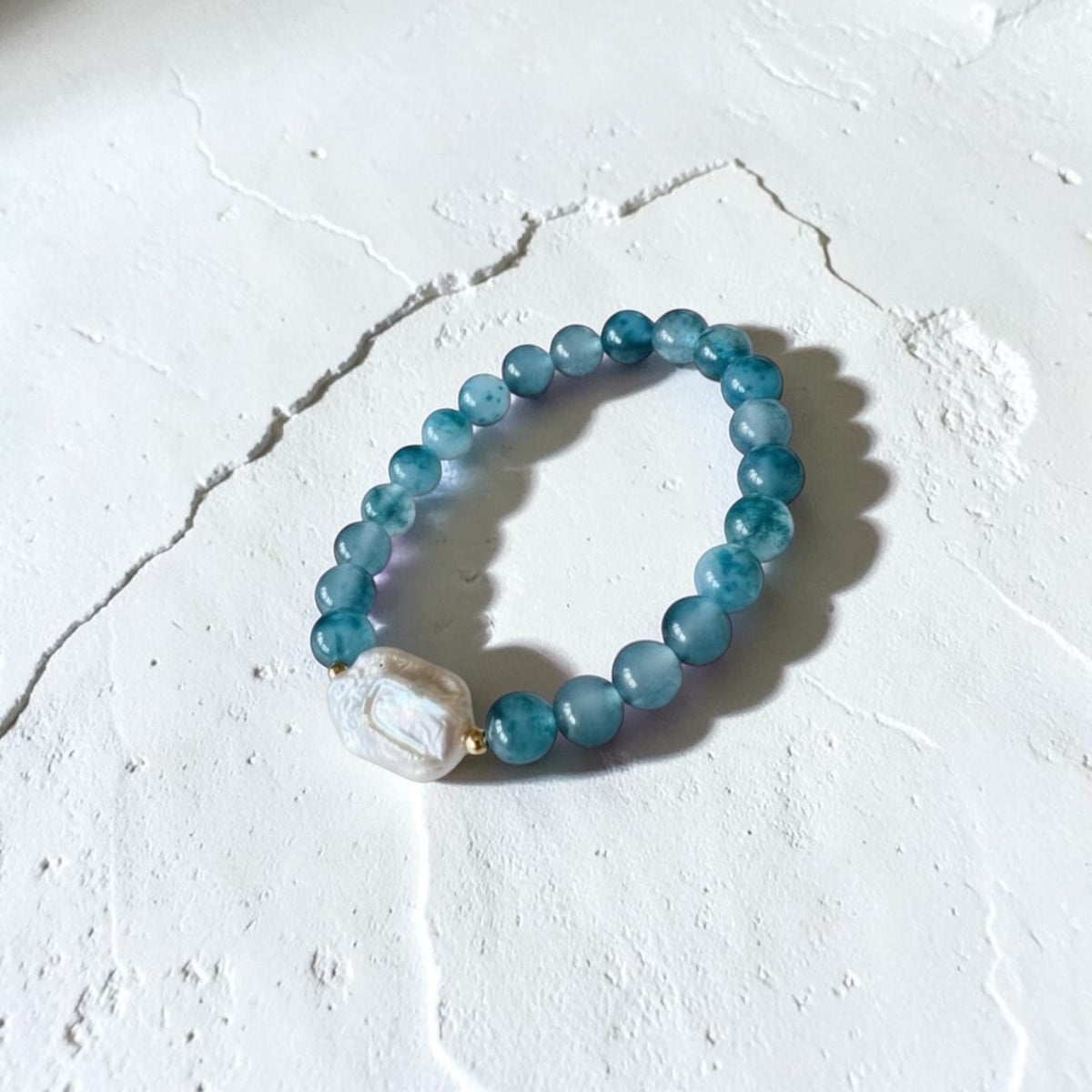 A beaded stretch bracelet featuring round light blue beads and a large, irregular white pearl-like accent bead, displayed on a textured white surface.