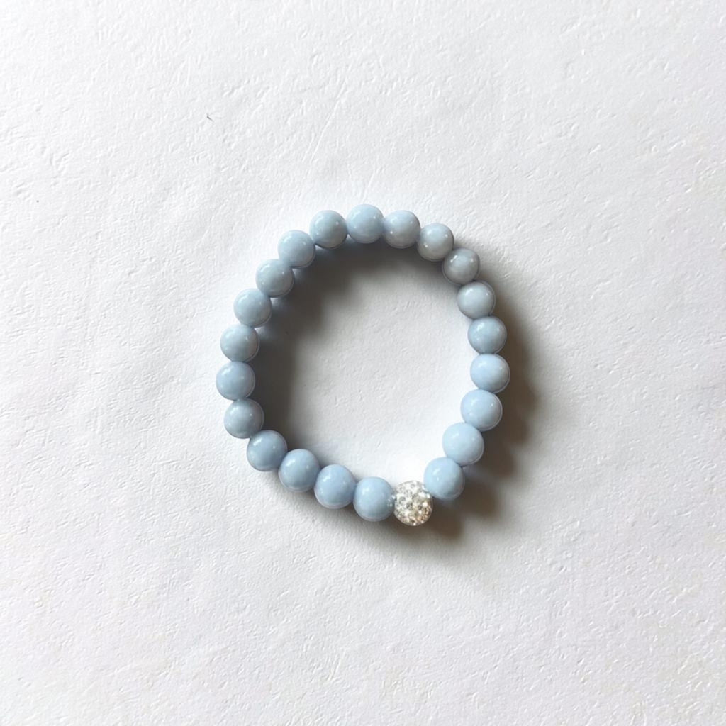 A light blue beaded stretch bracelet with a single sparkling clear rhinestone accent, displayed on a textured white surface.