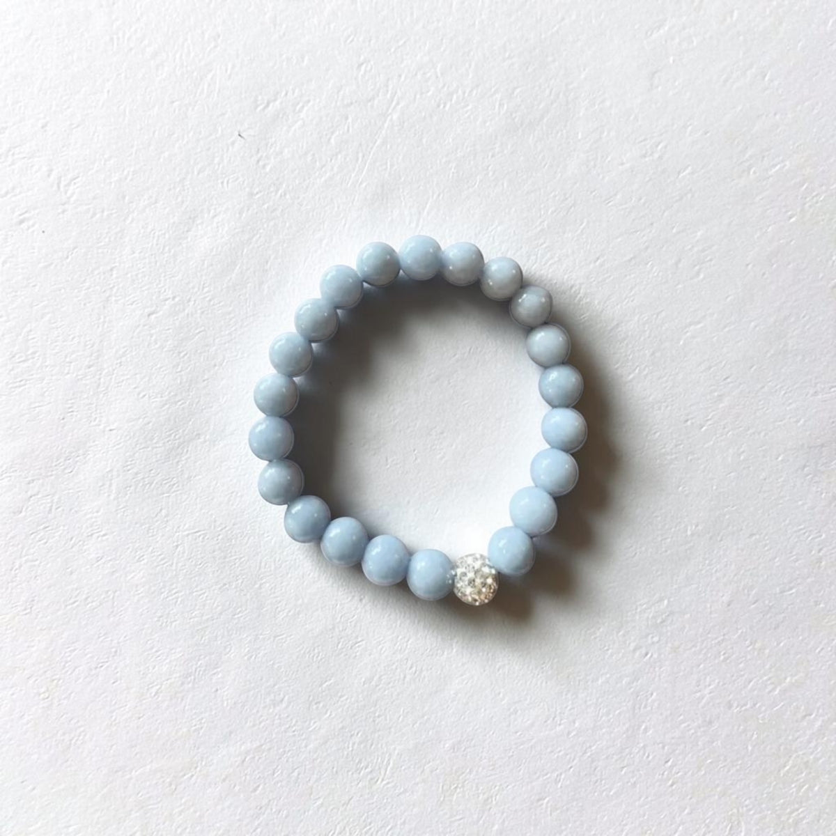 A light blue beaded stretch bracelet with a single sparkling clear rhinestone accent, displayed on a textured white surface.