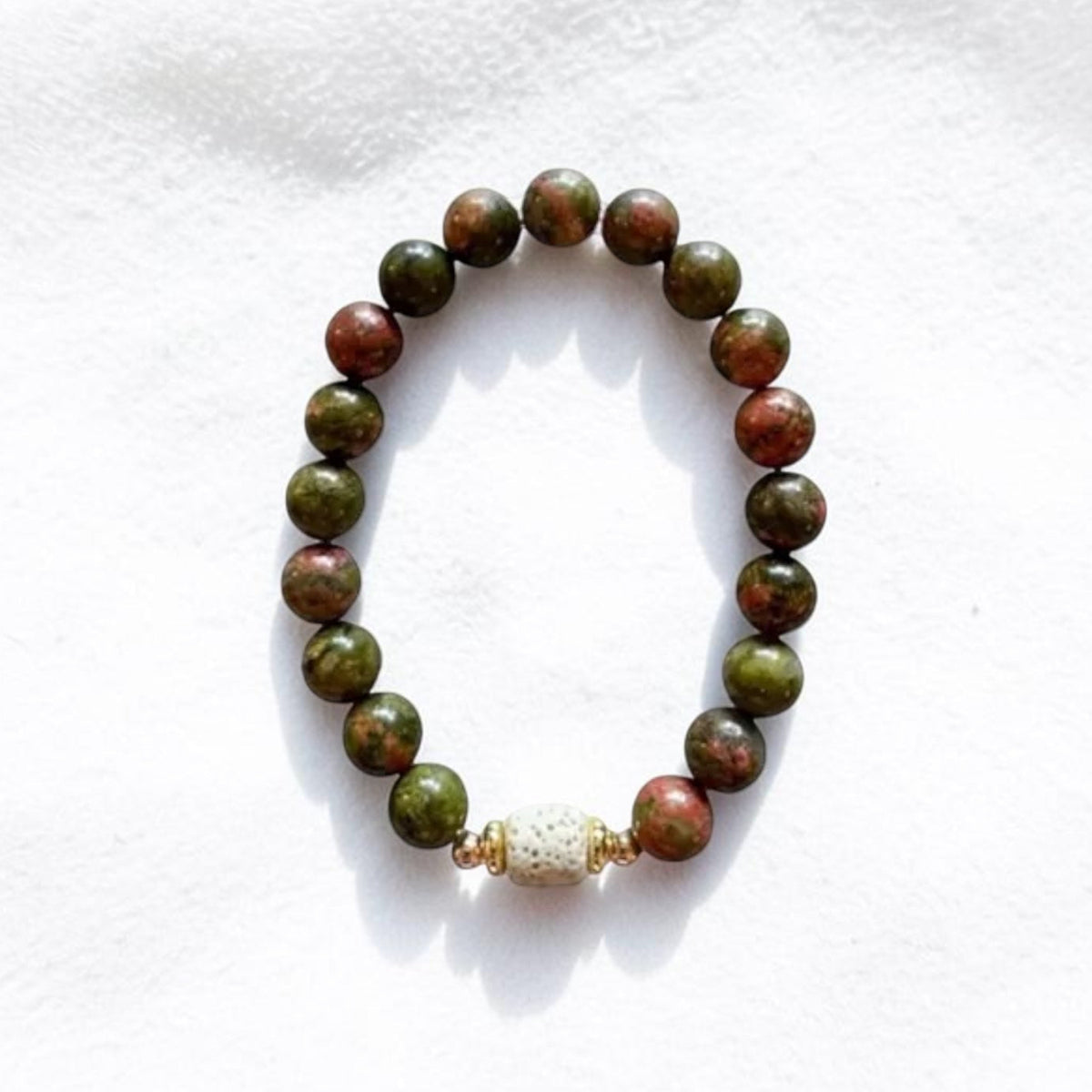 A beaded stretch bracelet made of round green and reddish-brown beads, with a small white textured spacer bead, displayed on a white textured fabric background.