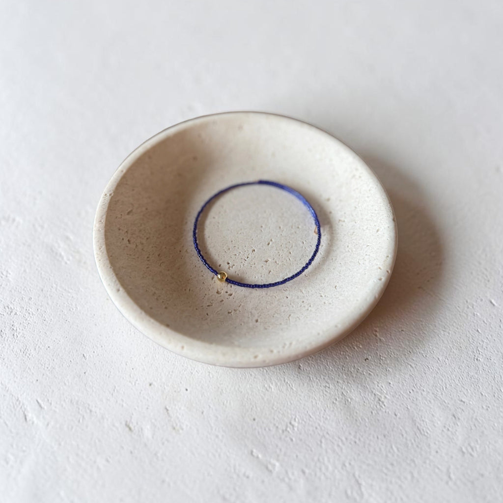 A small handmade ceramic dish with a speckled off-white glaze, featuring a thin purple blue wire bracelet inside its center, photographed on a textured light gray surface.