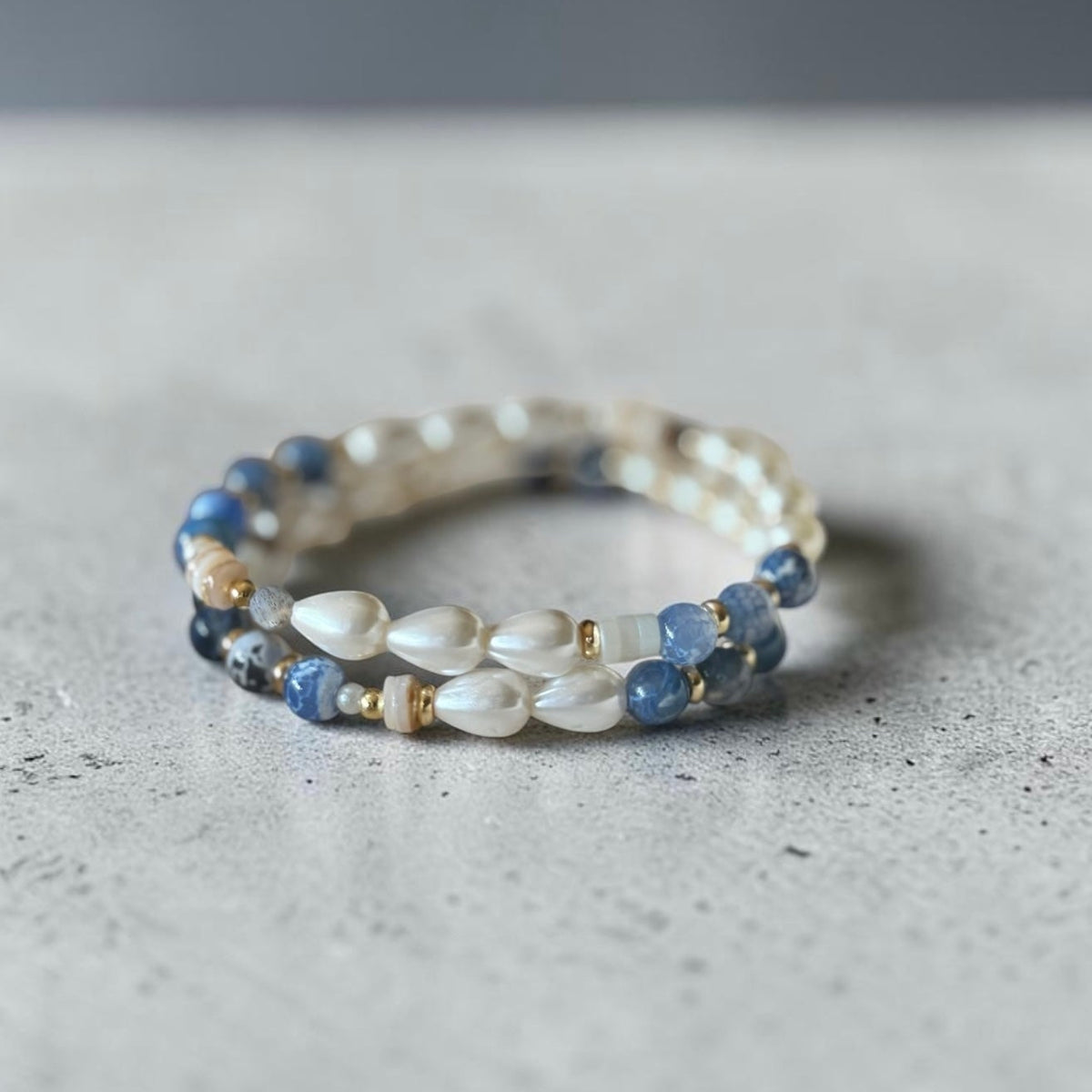 A stack of two beaded bracelet featuring white teardrop-shaped beads, round blue beads, and small gold spacer beads, resting on a textured gray surface.