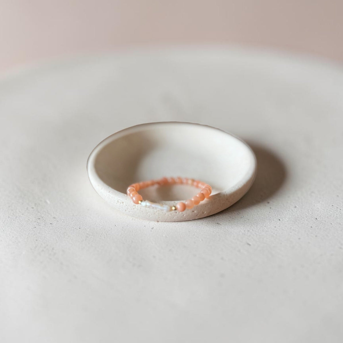 A delicate peach beaded bracelet with a small white bead accent, displayed inside a small, shallow, speckled ceramic dish on a textured light gray surface.