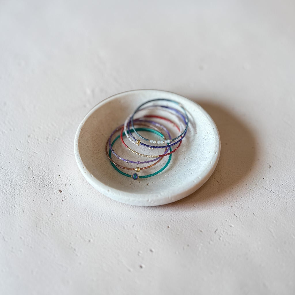 A small white ceramic dish holding several delicate multicolored beaded bracelets on a textured light beige surface.