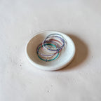 A small white ceramic dish holding several delicate multicolored beaded bracelets on a textured light beige surface.