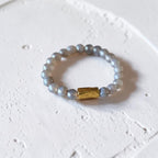 A beaded stretch ring featuring light gray-blue pearls and a textured gold rectangular accent bead, resting on a textured white surface.