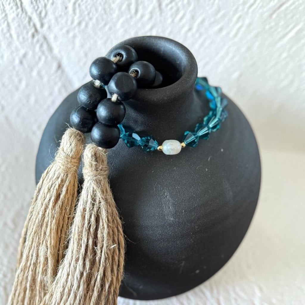 A beaded bracelet with teal faceted beads, a small pearl, displayed on a matte black ceramic vase against a textured off-white background.
