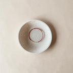 A round, speckled ceramic bowl in a warm off-white glaze, featuring a contrasting red painted circular accent inside the rim, photographed from directly above on a matching beige background.