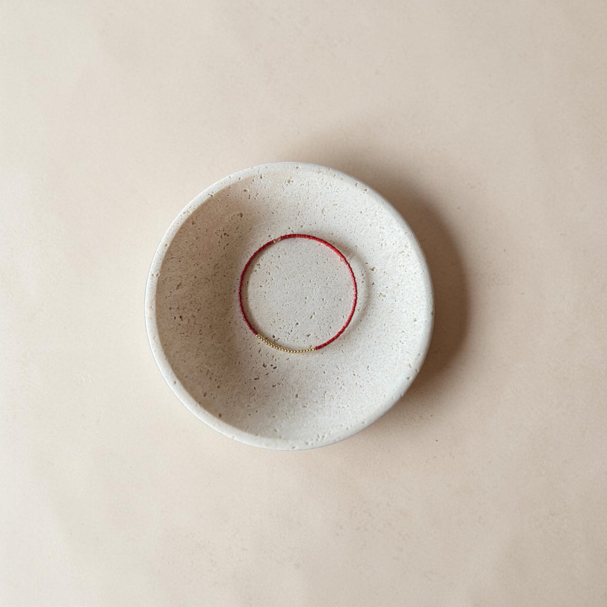 A round, speckled ceramic bowl in a warm off-white glaze, featuring a contrasting red painted circular accent inside the rim, photographed from directly above on a matching beige background.