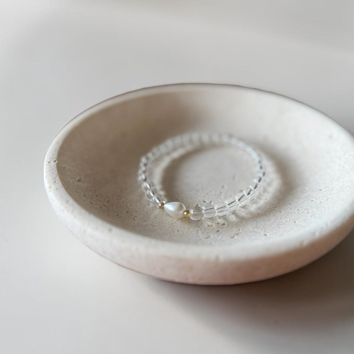 A delicate beaded bracelet with a small pearl and gold accent resting inside a round, textured ceramic dish on a soft neutral background.